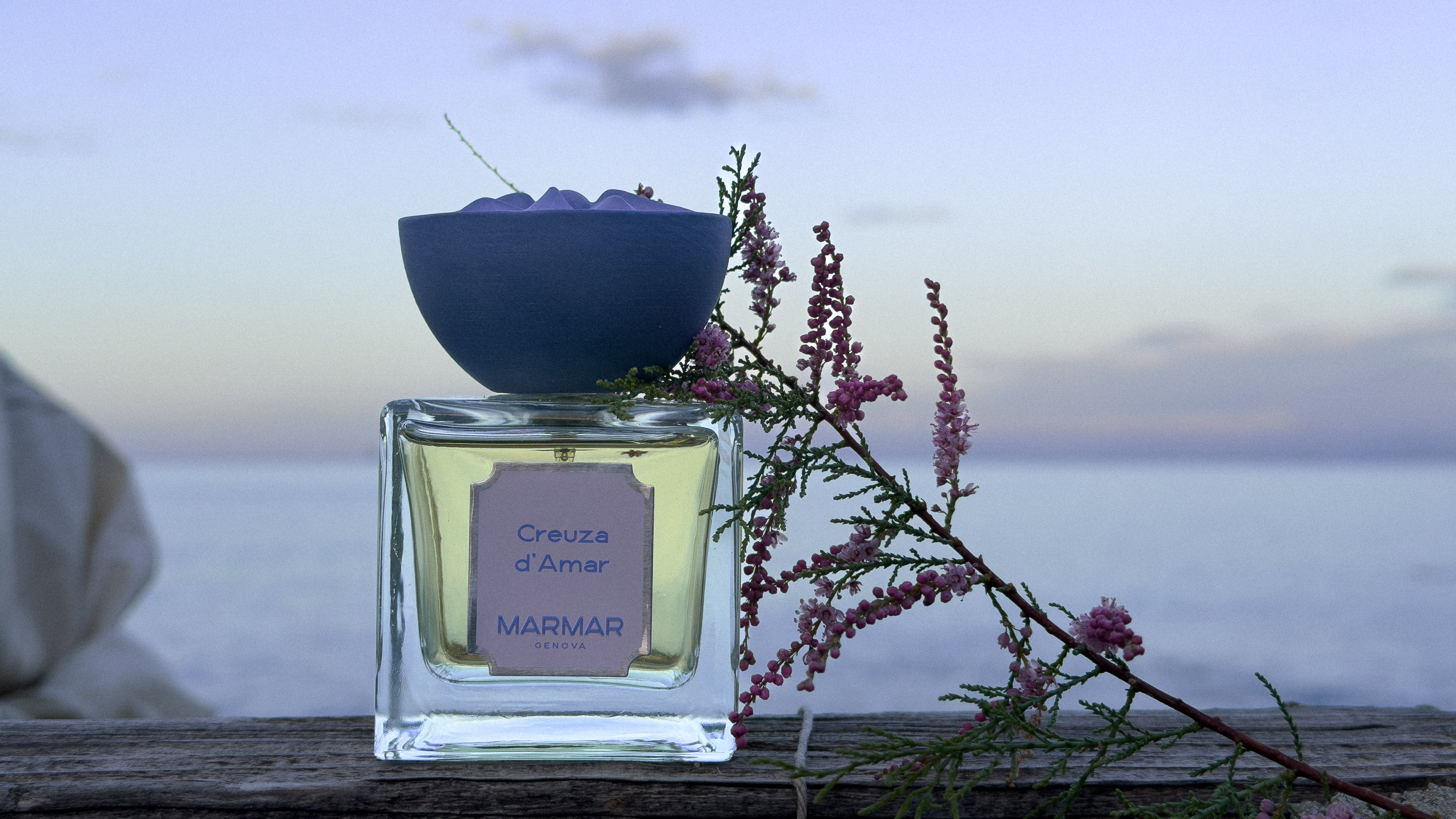 Still life of Creuza d'Amar eau de parfum by MarMar Genova shot against a sea backdrop at sunrise with a lilac floral branch.